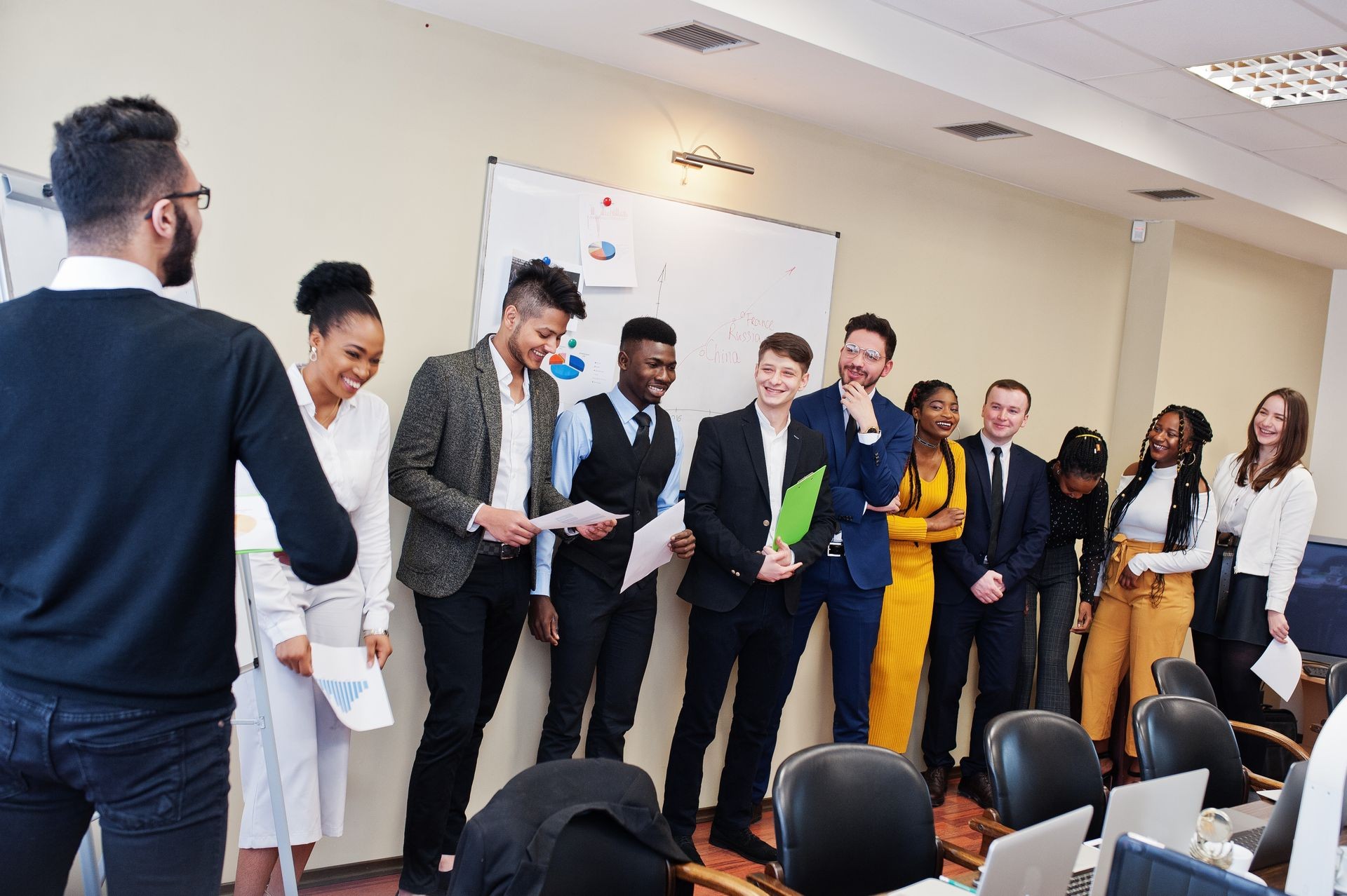 Mixed race business coach presenting report standing near whiteboard pointing on sales statistic shown on diagram and chart teach diverse company members gathered together in conference room.