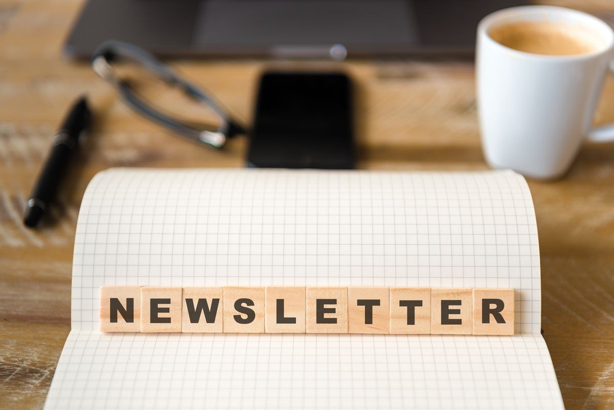 Closeup on notebook over wood table background, focus on wooden blocks with letters making Newsletter text. Concept image. Laptop, glasses, pen and mobile phone in defocused background